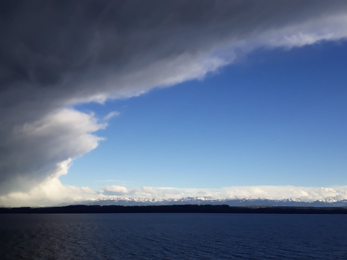 Vue sur le lac de Neuchâtel, nuages gris sur la gauche et ciel bleu sur la droite. Alpes enneigées dans le fond